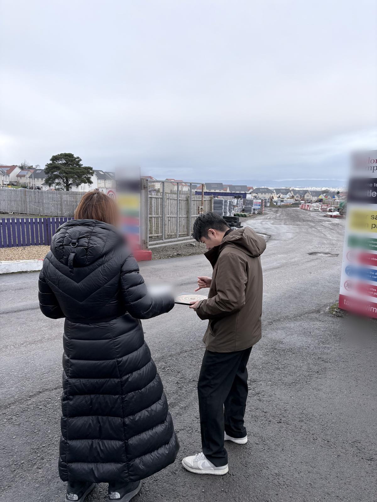 Daniel Siew holding a luopan Feng Shui compass at the entrance to a residential development site in Glasgow, Scotland, with houses under construction behind.