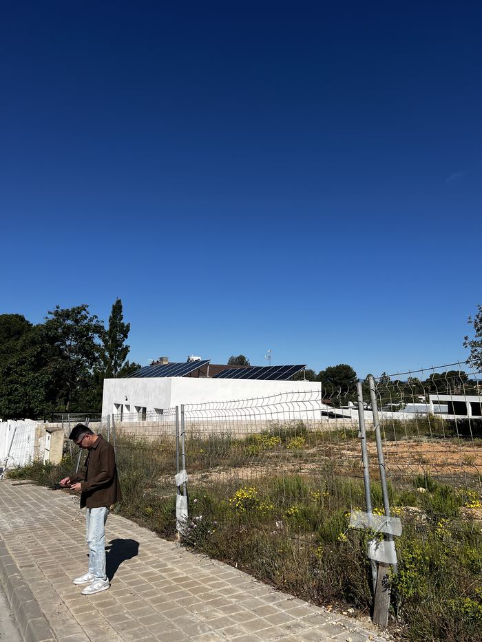 Daniel Siew standing at the fenced boundary of a vacant plot in Valencia, Spain, taking a reading, with neighbouring houses behind the site.